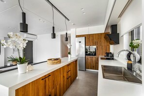 A modern kitchen with a central island, wooden cabinets, stainless steel appliances, pendant lighting, and a view into the dining area. - Kamelia Luxury Villa 1 (Hersonissos)