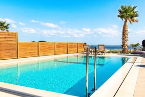 A swimming pool with a wooden fence, lounge chairs, and a palm tree. - Kamelia Luxury Villa 1 (Hersonissos)