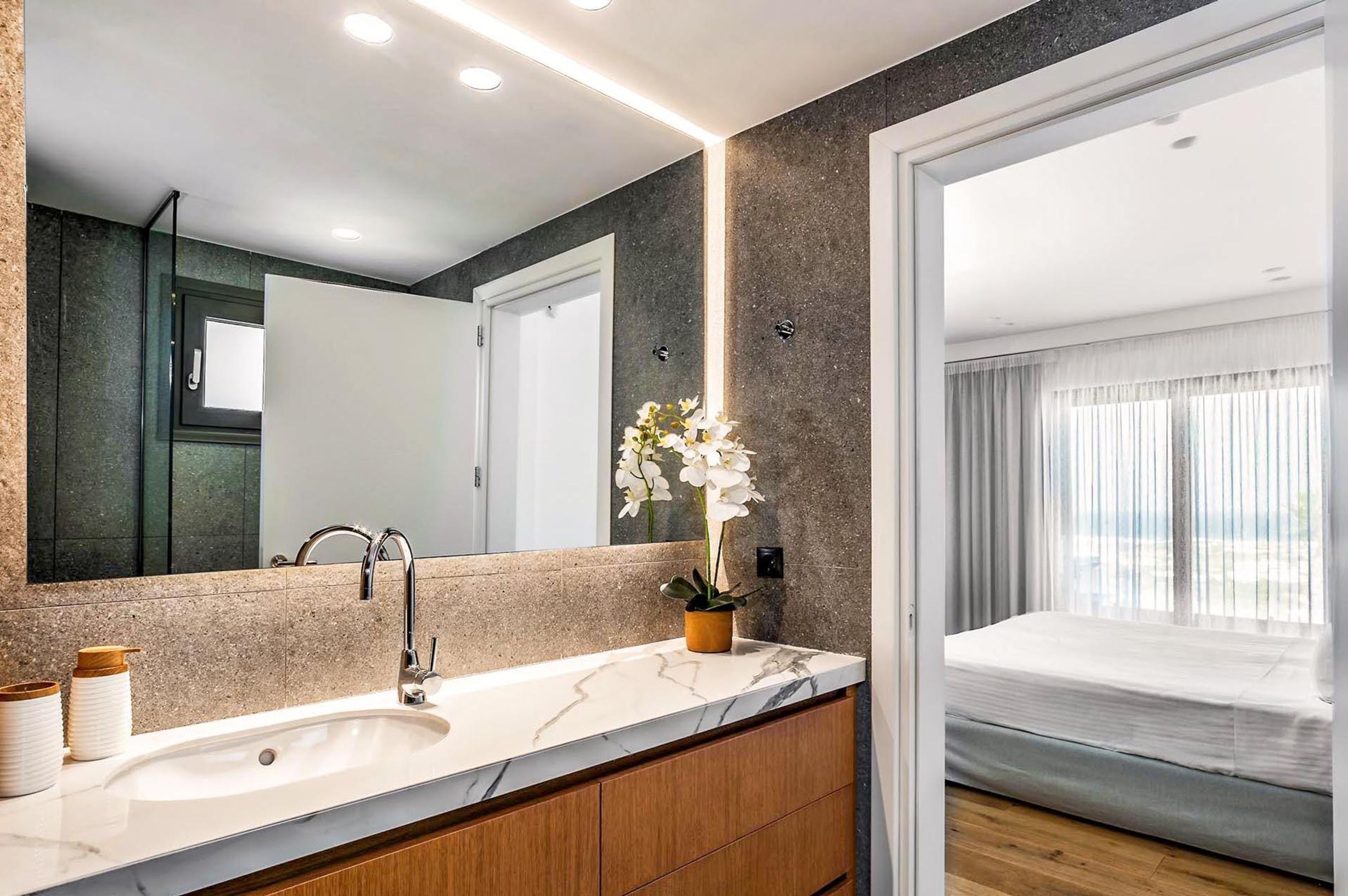 A modern bathroom with a marble sink, wooden cabinets, a large mirror, and a potted orchid.