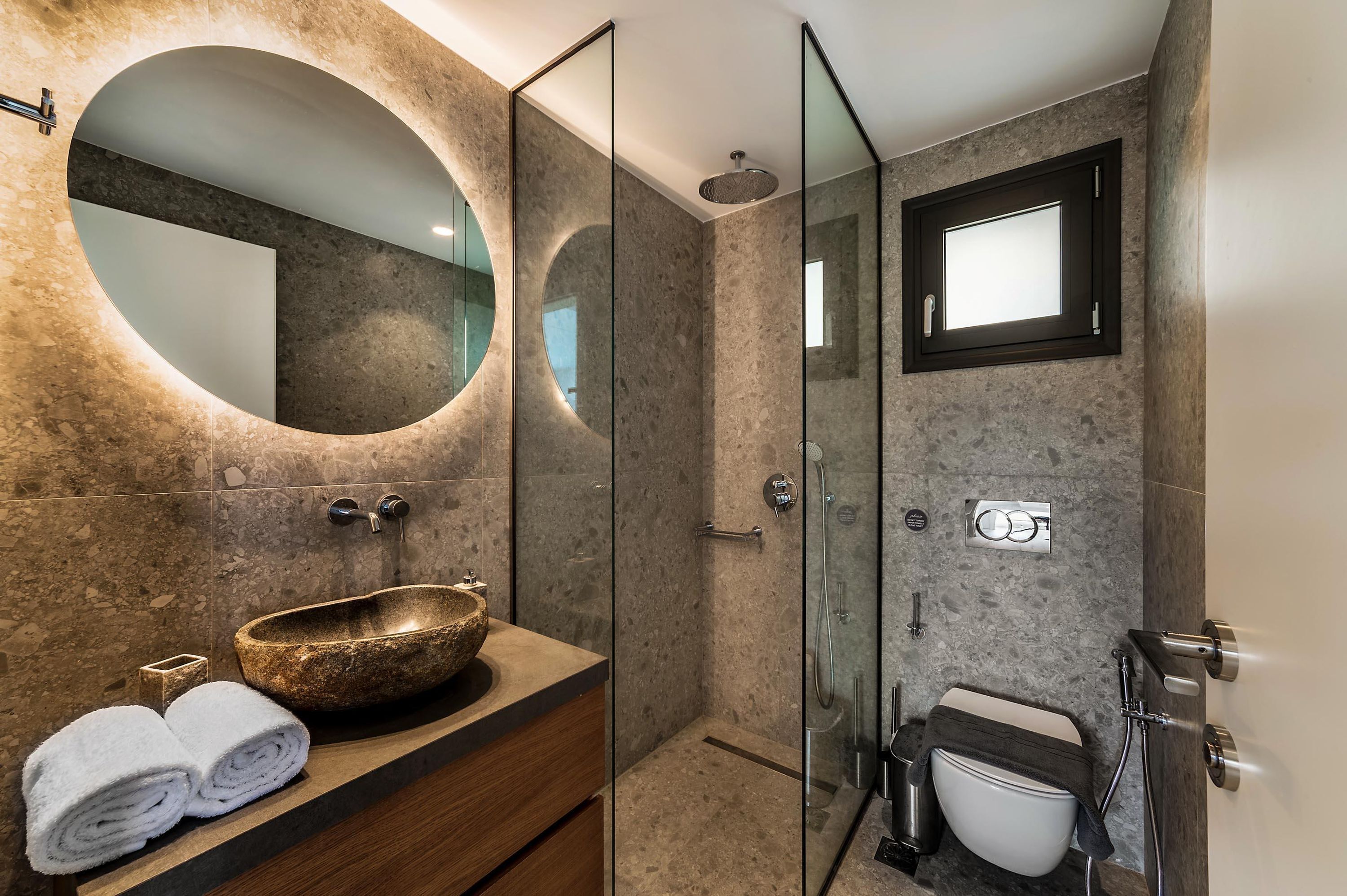 A modern bathroom with a round mirror, a stone sink, and a glass-enclosed shower.