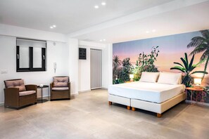 Une chambre moderne avec un grand lit, deux fauteuils en osier, une petite table, une prise électrique fixée au mur et une porte.
