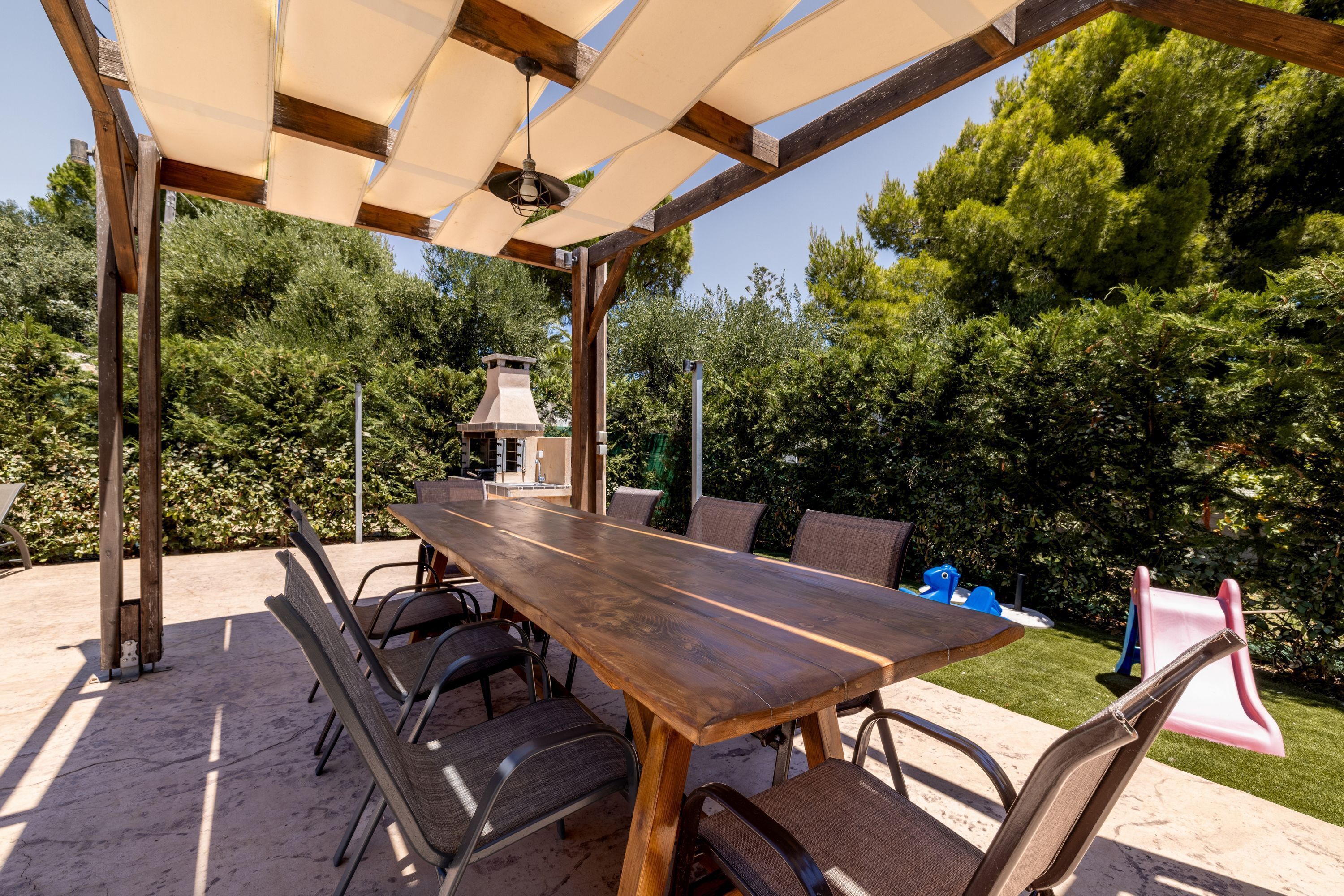 Un patio al aire libre con mesa y sillas de madera, rodeado por una pérgola con una lámpara.
