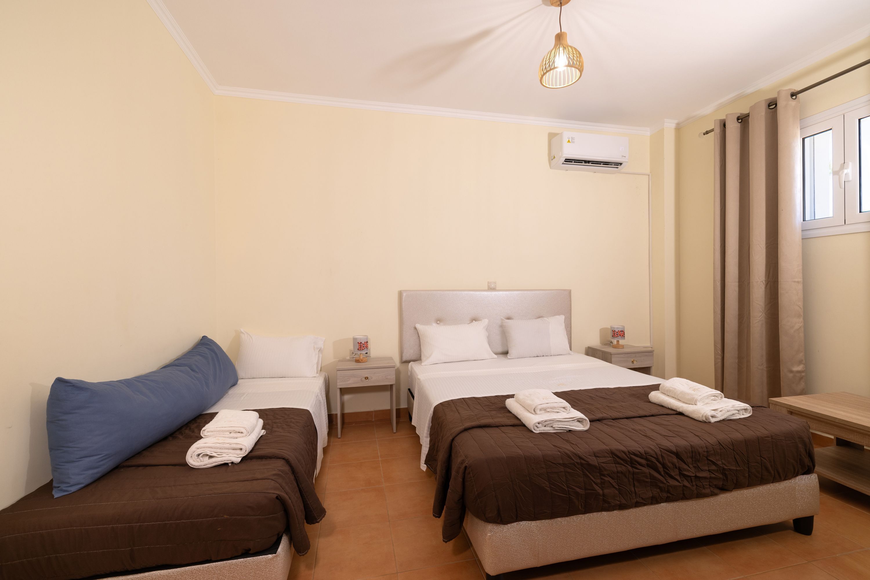 A hotel room with two beds, a small bedside table with a lamp, a wall-mounted air conditioner, and a window with curtains.