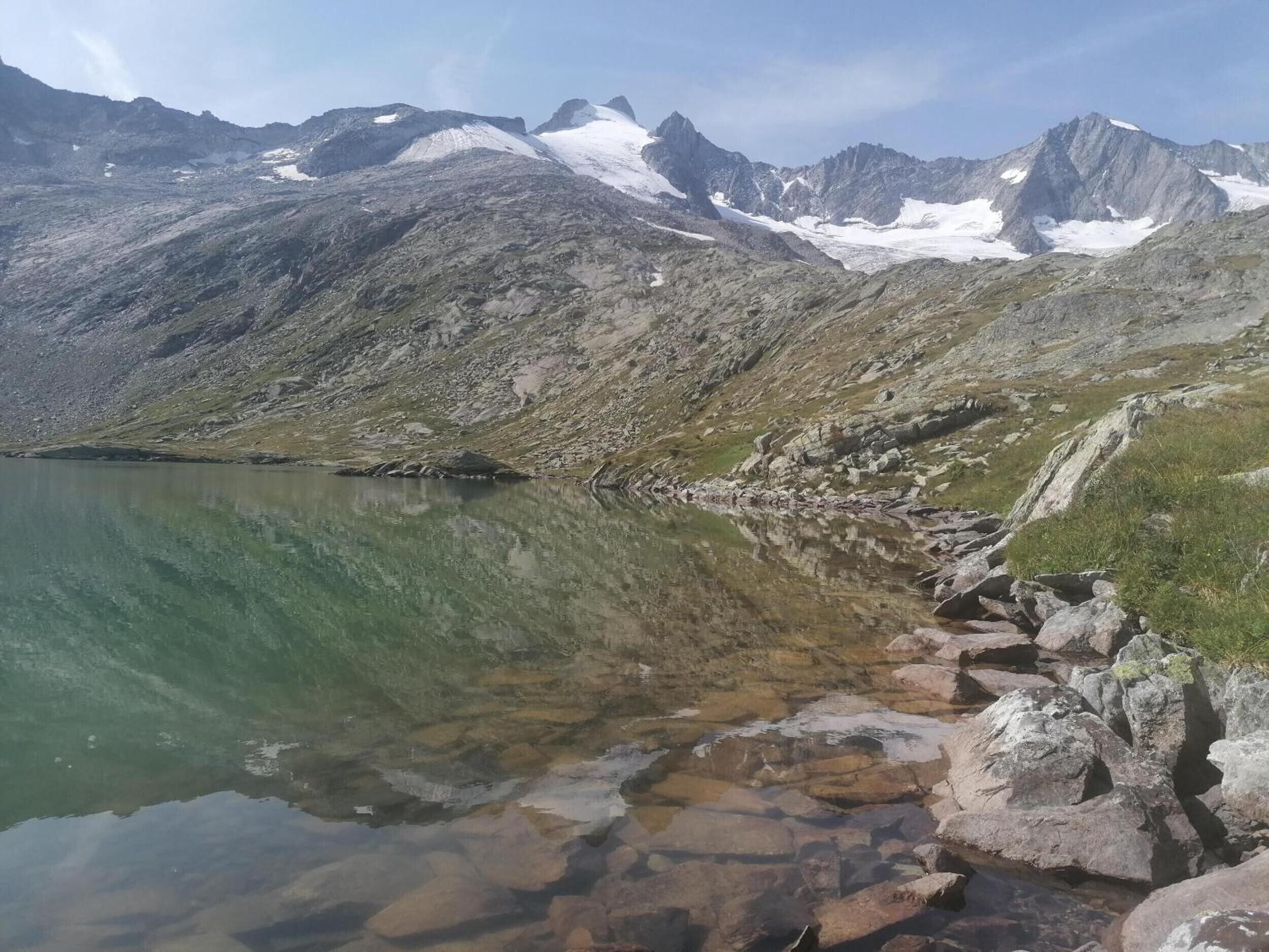 Eine Berglandschaft mit einem klaren See, felsigem Ufer und schneebedeckten Gipfeln.