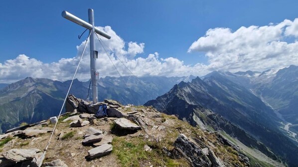 A mountain peak with a cross, rocky terrain, and a clear view of distant mountains.