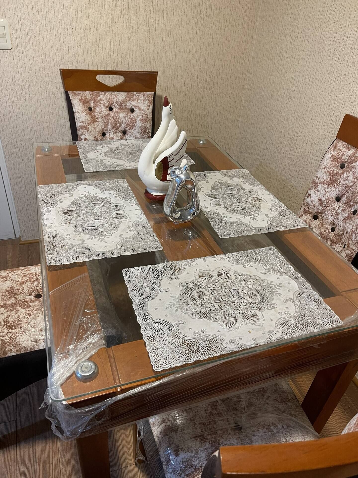 A dining table with four chairs, each with a swan-shaped cushion. The table is covered with a glass top and four square placemats with intricate lace patterns.