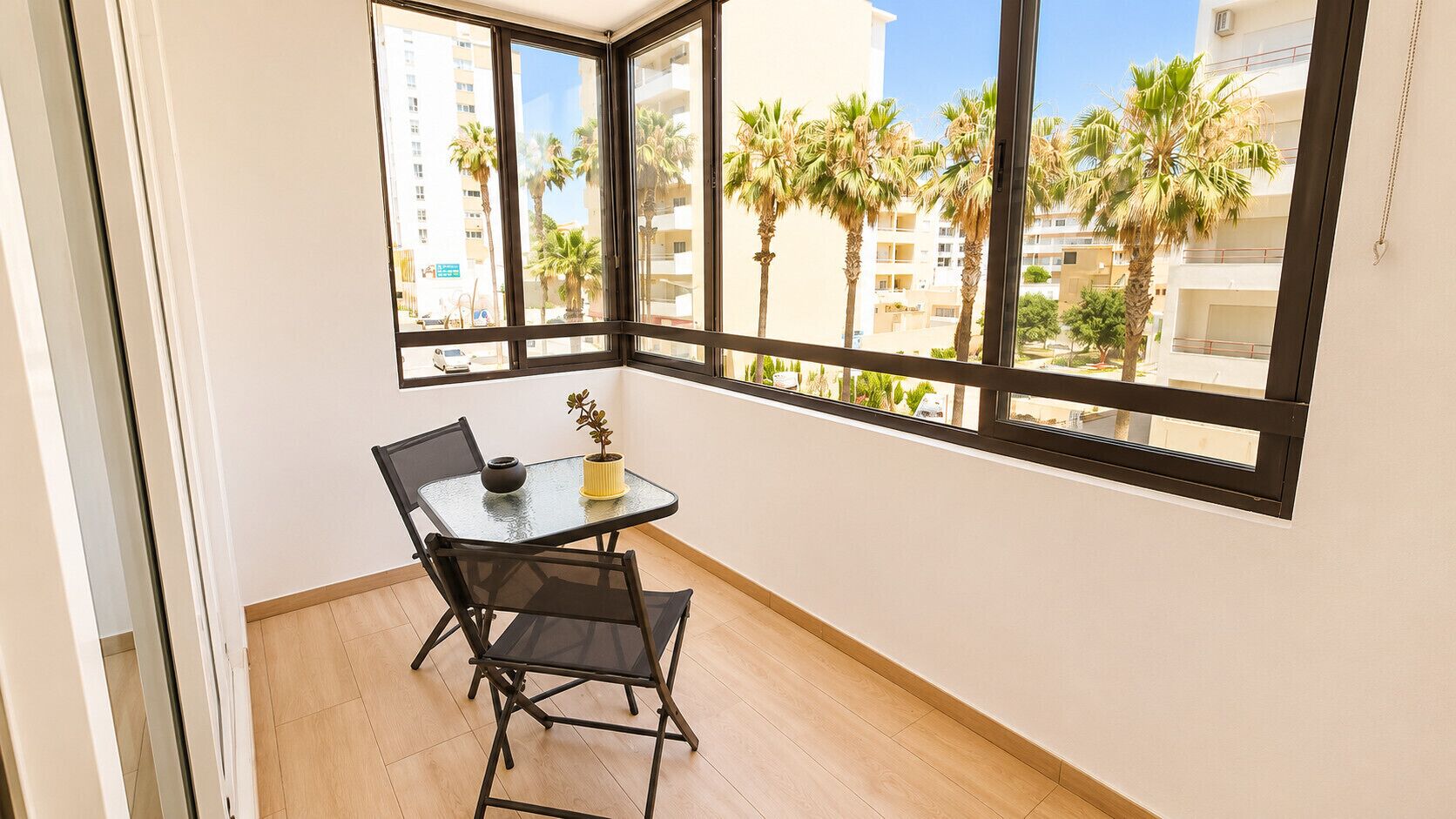 A balcony with a table and chairs, a potted plant, and a view of palm trees and buildings.