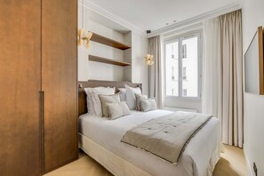 A bedroom with a large bed, wooden wardrobe, built-in shelves, a window with curtains, and a wall-mounted light fixture. - Superbe Apartment Saint Honoré Elysée Palace (Paris)