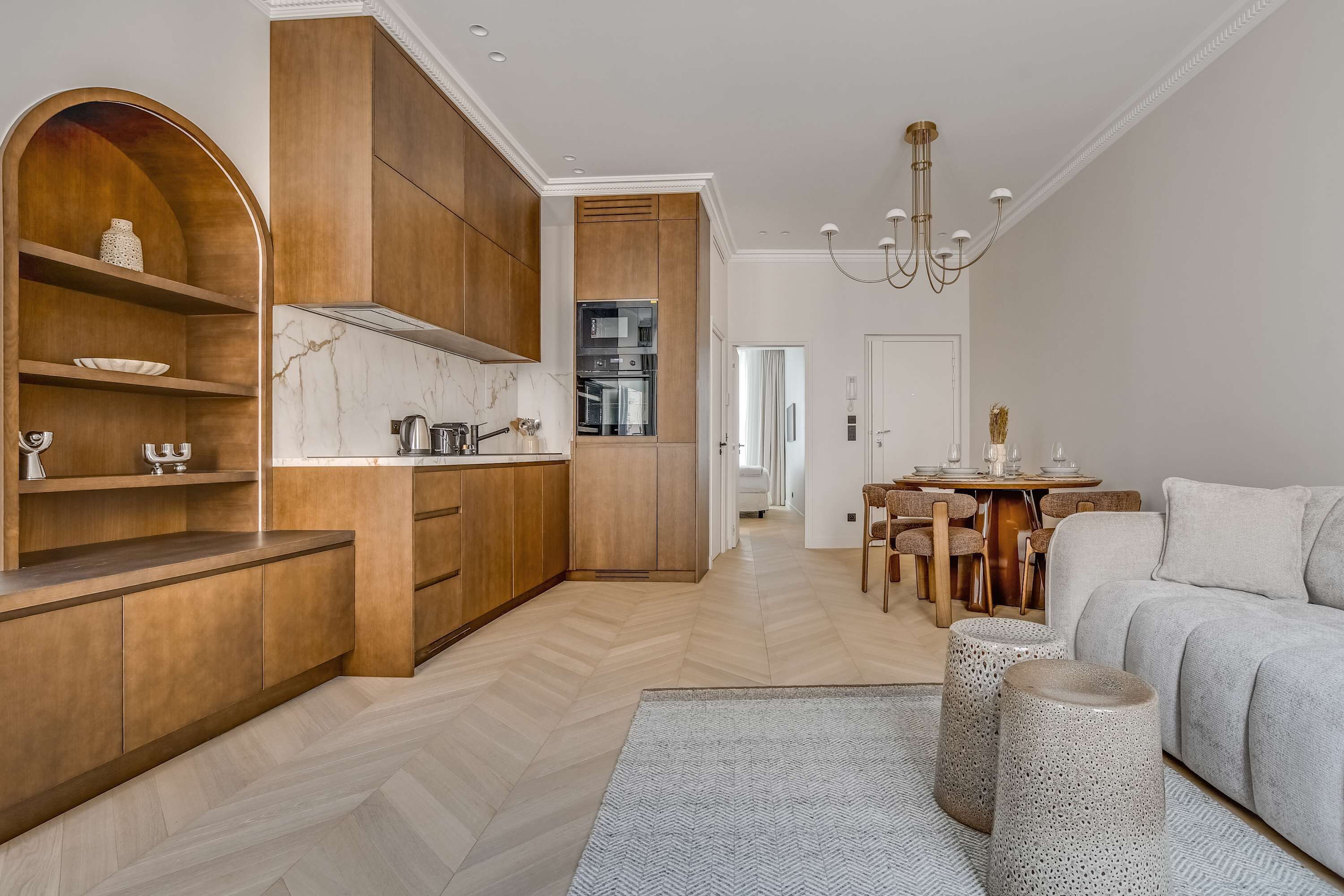 A modern kitchen with wooden cabinets and a marble countertop, leading to a dining area with a chandelier and a sofa.
