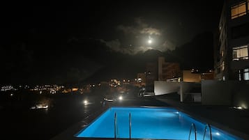 Zona de piscina nocturna con vista a un paisaje urbano y una montaña al fondo.