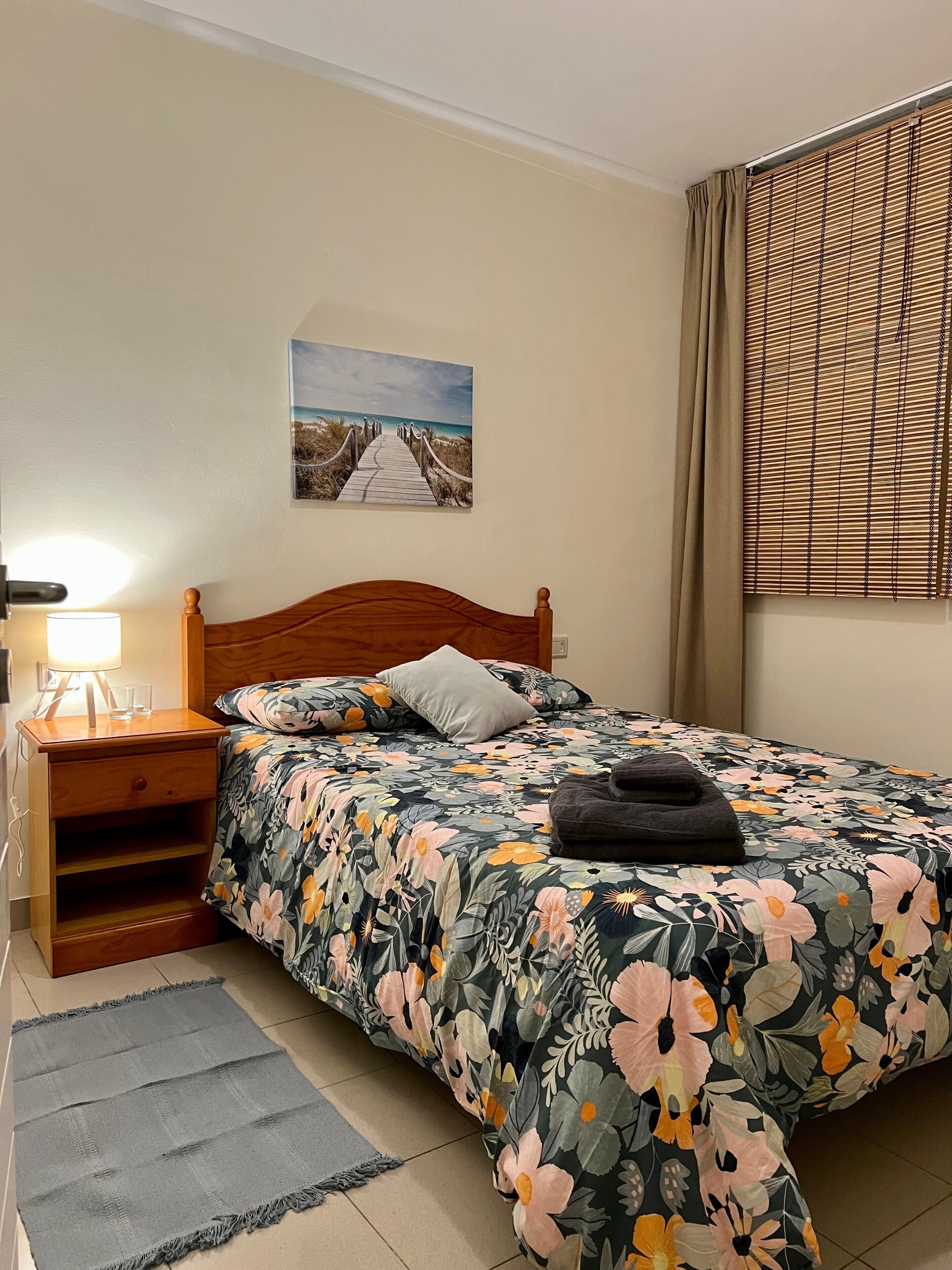 A bedroom with a wooden headboard, a bed with floral bedding, a nightstand with a lamp, a picture on the wall, and a window with blinds.