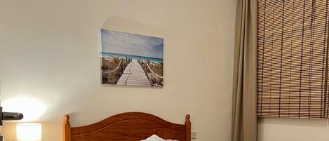 A bedroom with a wooden headboard, a bed with floral bedding, a nightstand with a lamp, a picture on the wall, and a window with blinds.