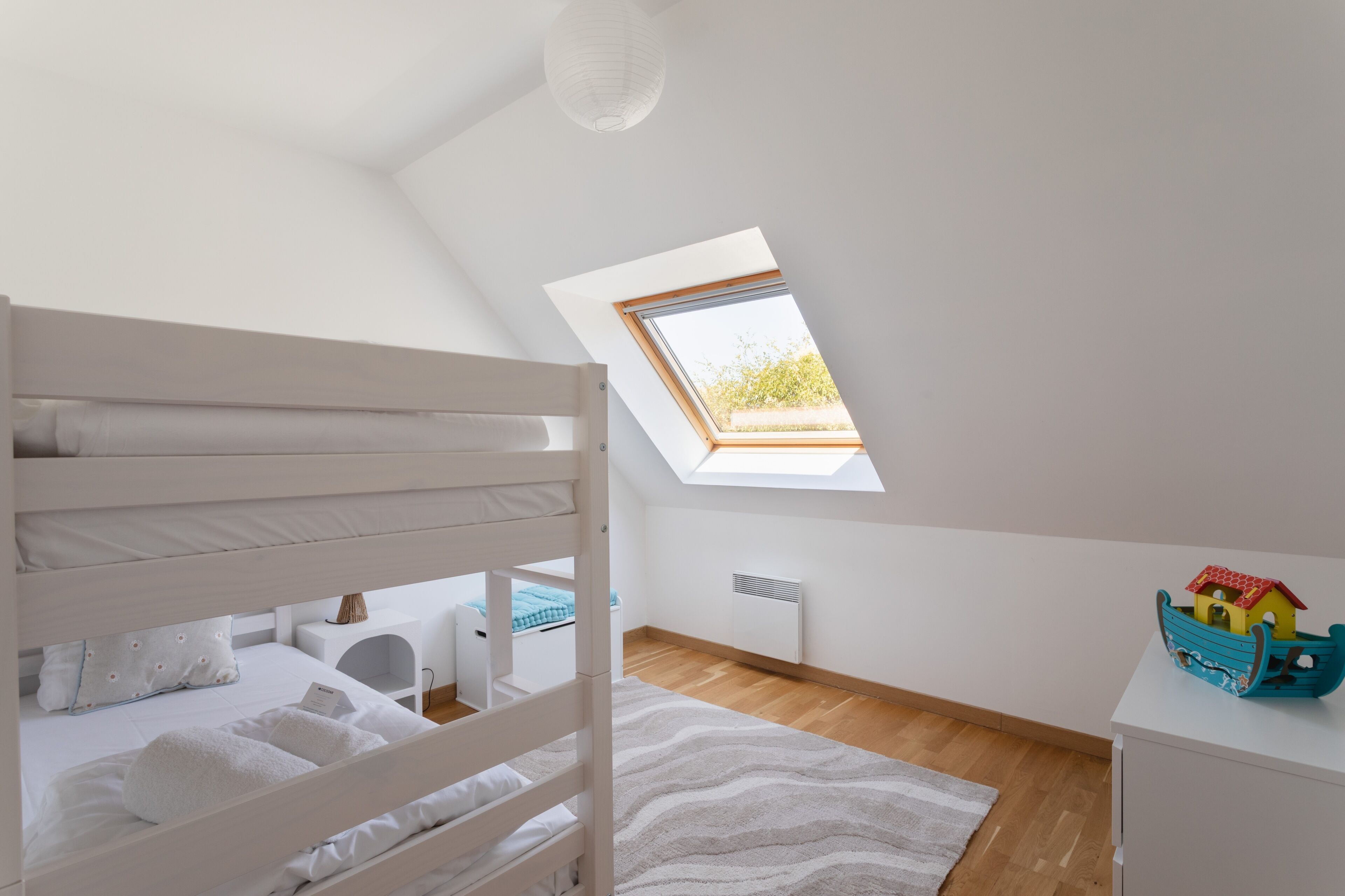 A child's bunk bed with a skylight above, a small white dresser, and a toy house on the dresser.