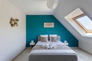 Ein Schlafzimmer mit schrägem Dach, einem Oberlicht, einem Bett mit weißer Bettwäsche, zwei gemusterten Kissen, einem gerahmten Kunstwerk und einem kleinen Nachttisch mit einem Glasobjekt.
