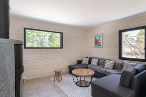 Ein modernes Wohnzimmer mit einem grauen Sofa, einem runden Holztisch und einem kleinen Hocker. Zwei Fenster sorgen für natürliches Licht und bieten einen Blick ins Freie.