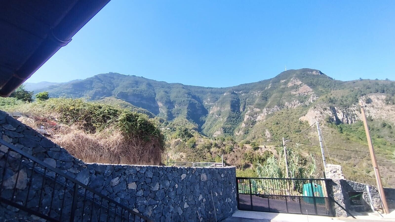 Un muro de piedra con barandilla metálica, vista de un paisaje montañoso y cielo despejado.