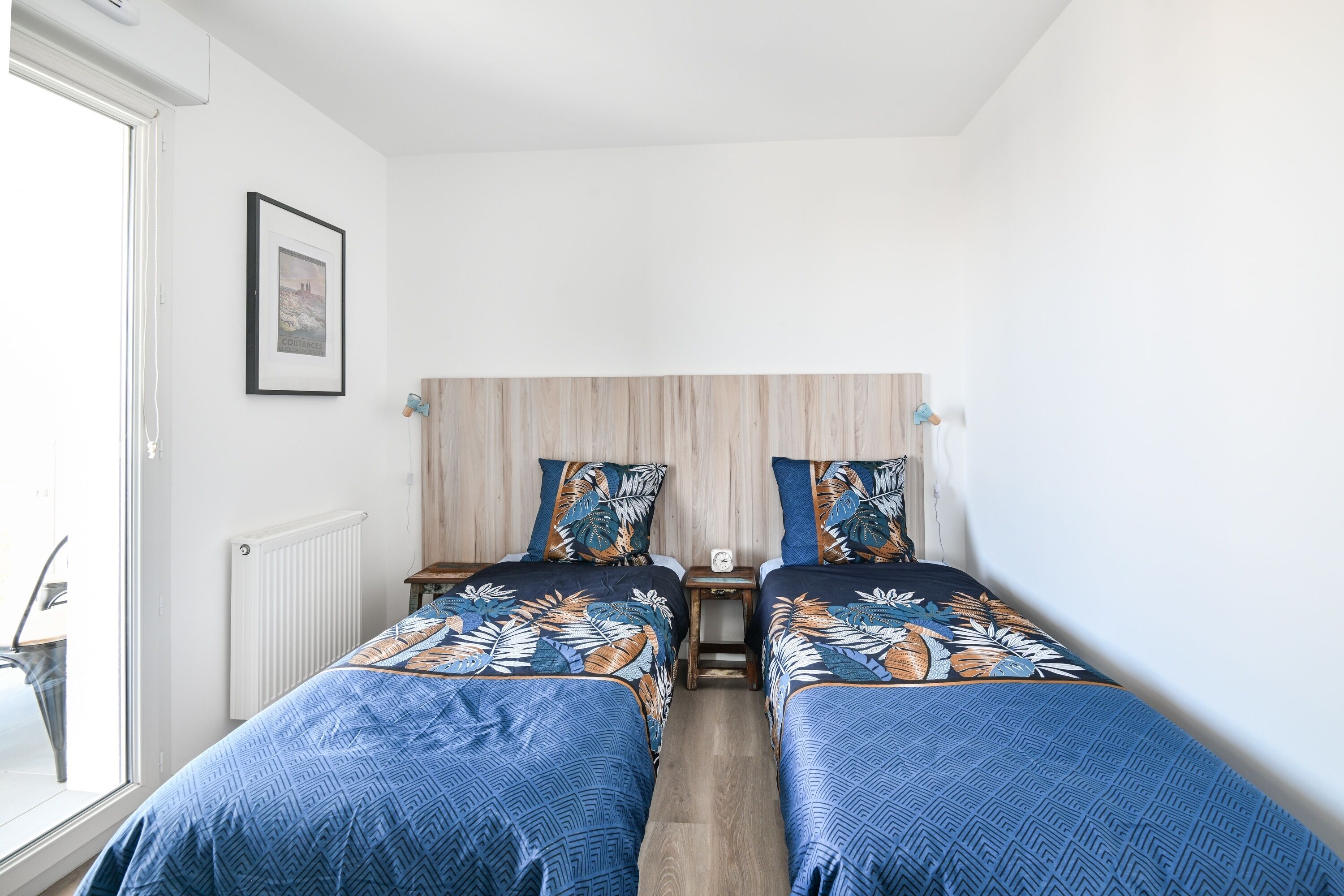 Quarto com duas camas, cabeceira de madeira, quadro na parede e janela à esquerda.