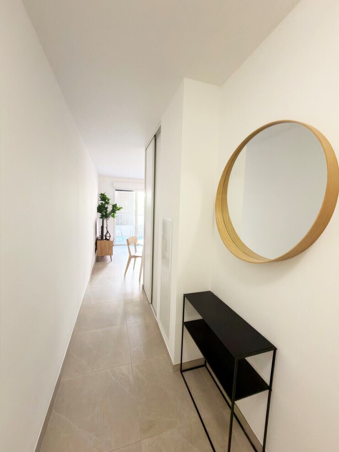 Un couloir avec un grand miroir rond accroché au mur, une console noire et une plante visible au loin.