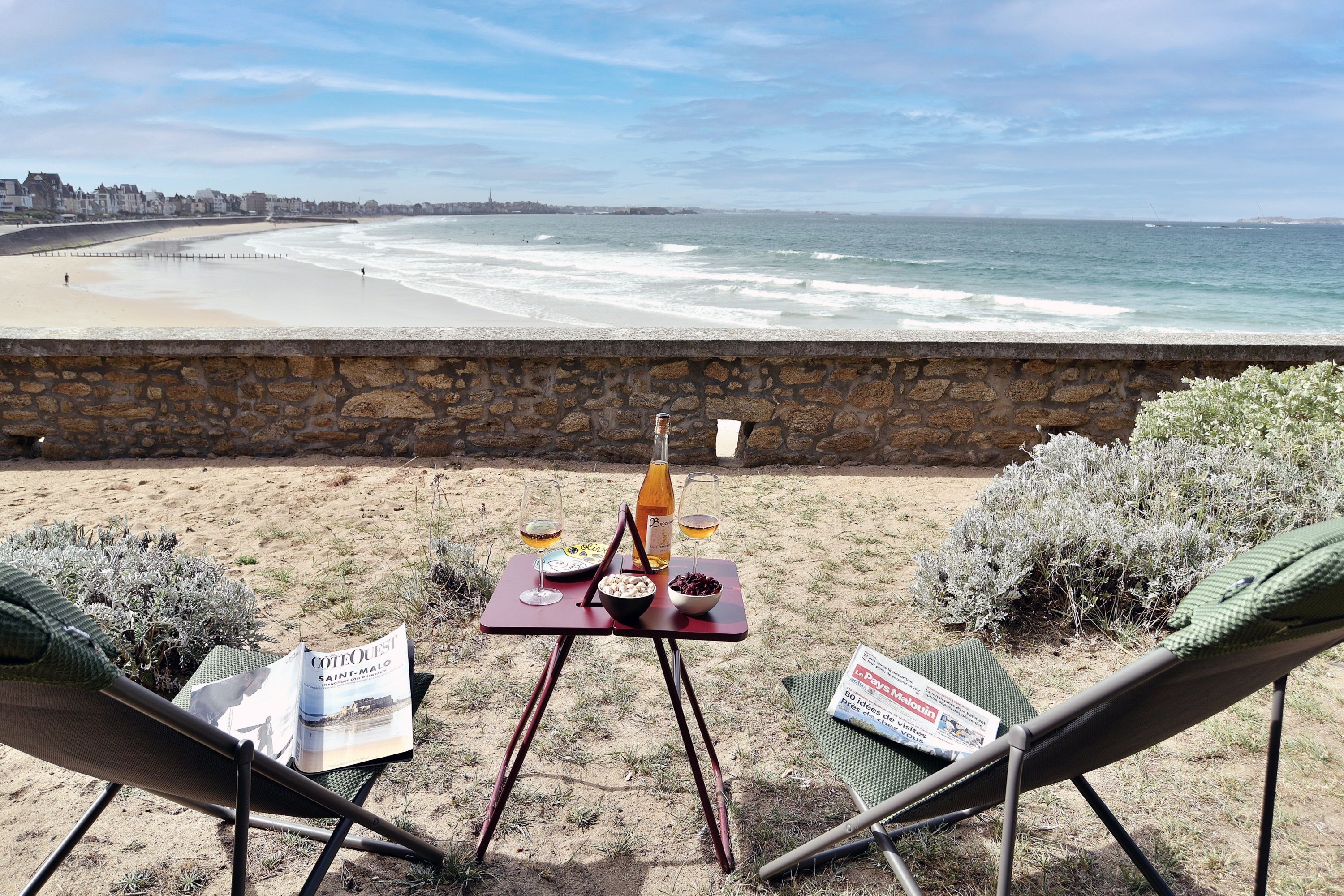 A beachside table with a bottle of wine, a glass, and a bowl of nuts, set against a backdrop of a sandy beach and ocean waves.