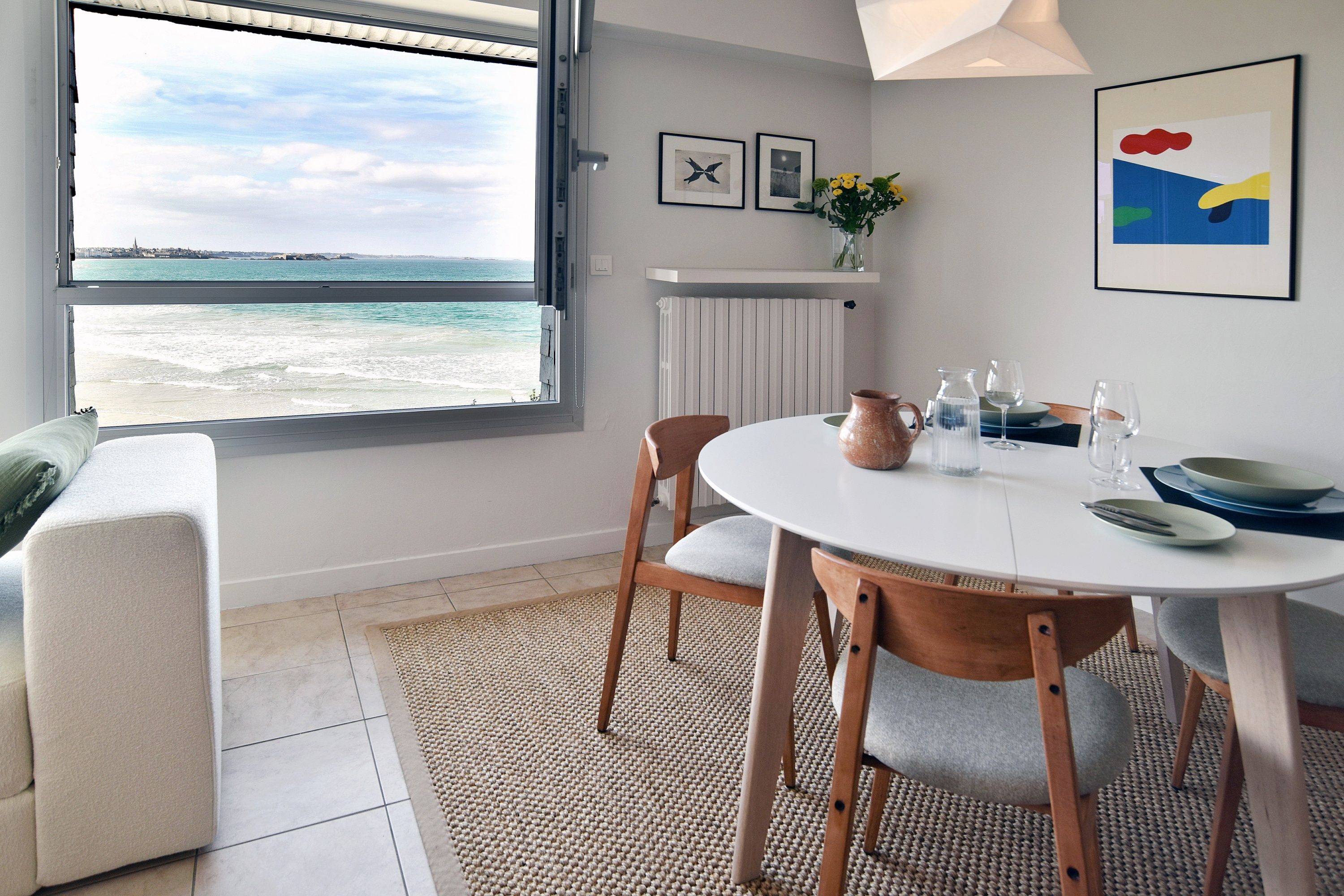 A dining area with a view of the sea, a white table, wooden chairs, and a vase with flowers.