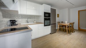 A modern kitchen with white cabinetry, a built-in oven, and a dining area with wooden chairs and a table. - Le Cèdre - Studio avec Parking (Janvry)