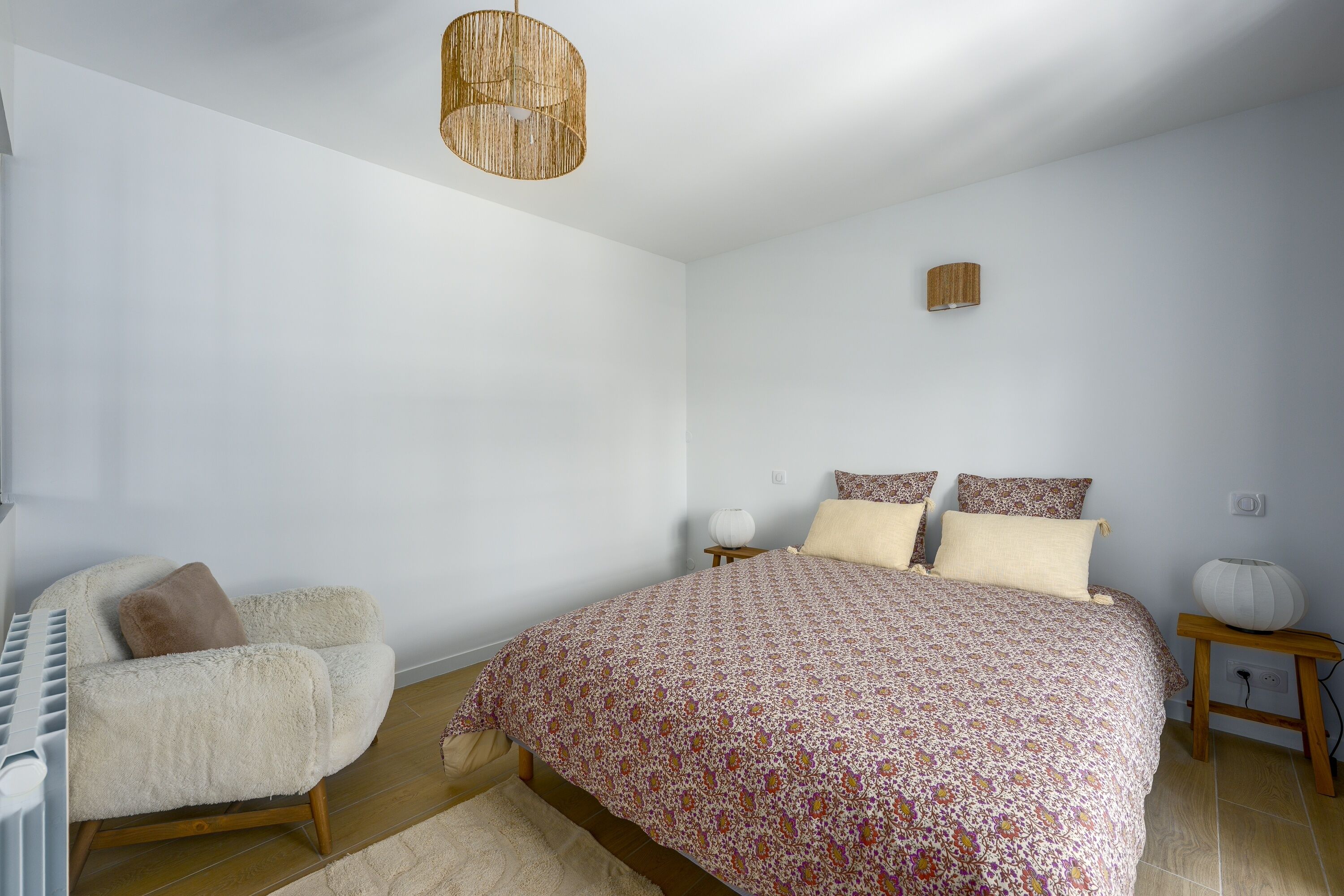 Un dormitorio con cama, una silla, mesita de noche, lámpara y una lámpara colgante.