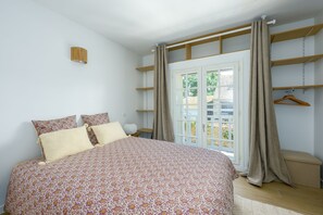 Un dormitori amb llit, finestra amb cortines, prestatge de fusta i una estant per a roba penjada.