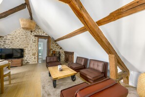 A cozy loft bedroom with a stone wall, wooden beams, a television, a sofa, and a coffee table. - Le Chêne - Studio avec Parking (Janvry)