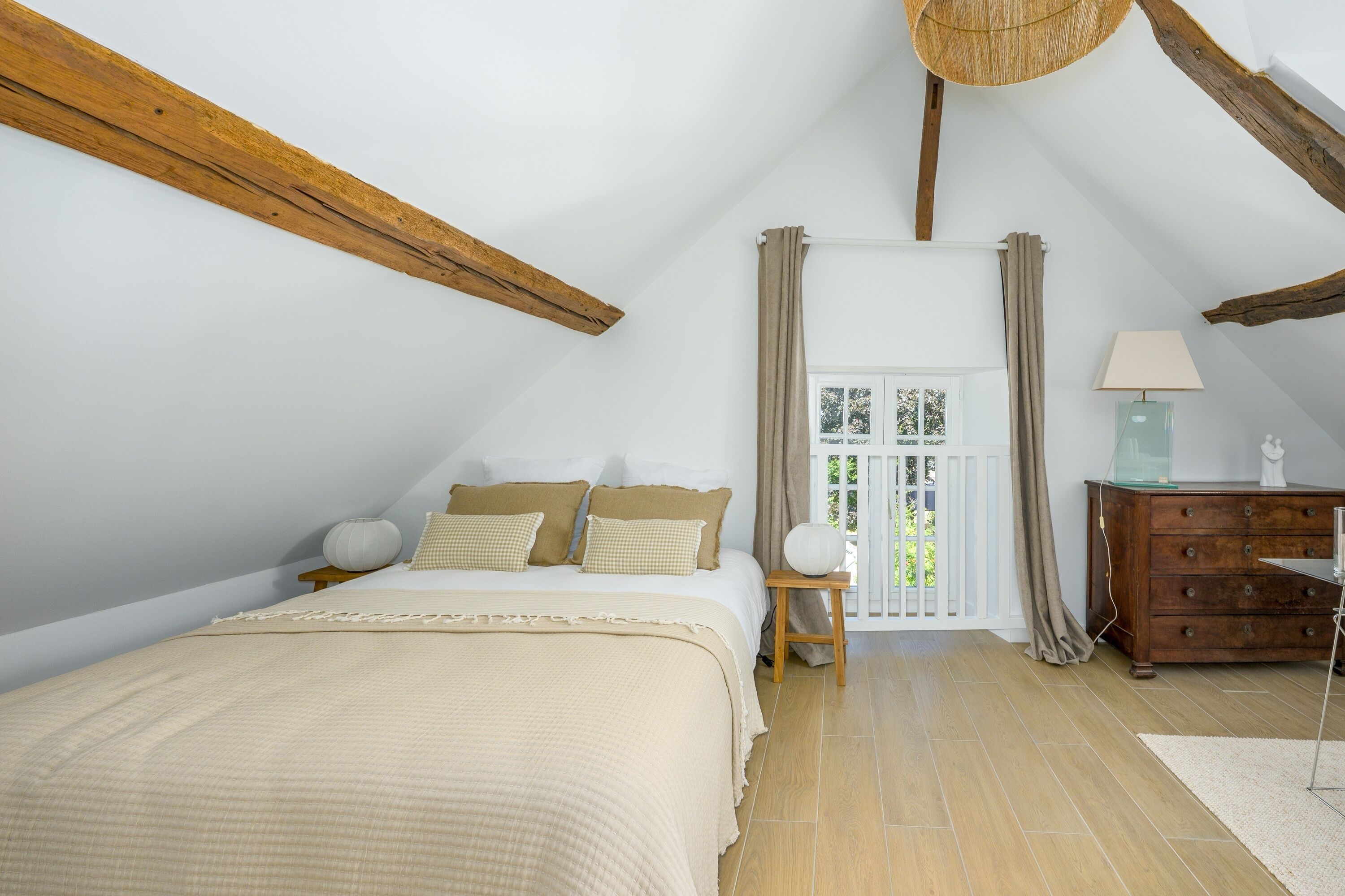 A bedroom with a sloped ceiling, a bed with a beige blanket, a bedside table with a lamp, and a dresser.