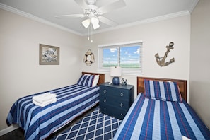 Une chambre avec deux lits, un ventilateur de plafond, une fenêtre avec des stores et des éléments de décoration murale sur le thème de la marine.