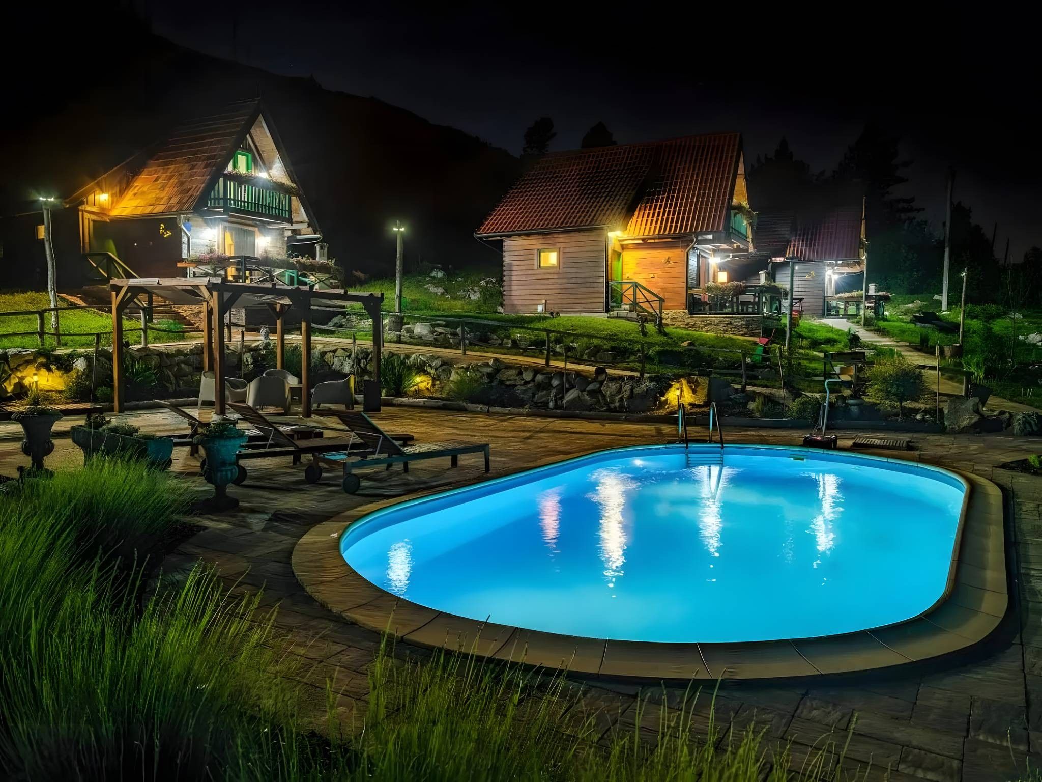 A swimming pool with lounge chairs and a gazebo, surrounded by a landscaped garden and a wooden building with a red roof.