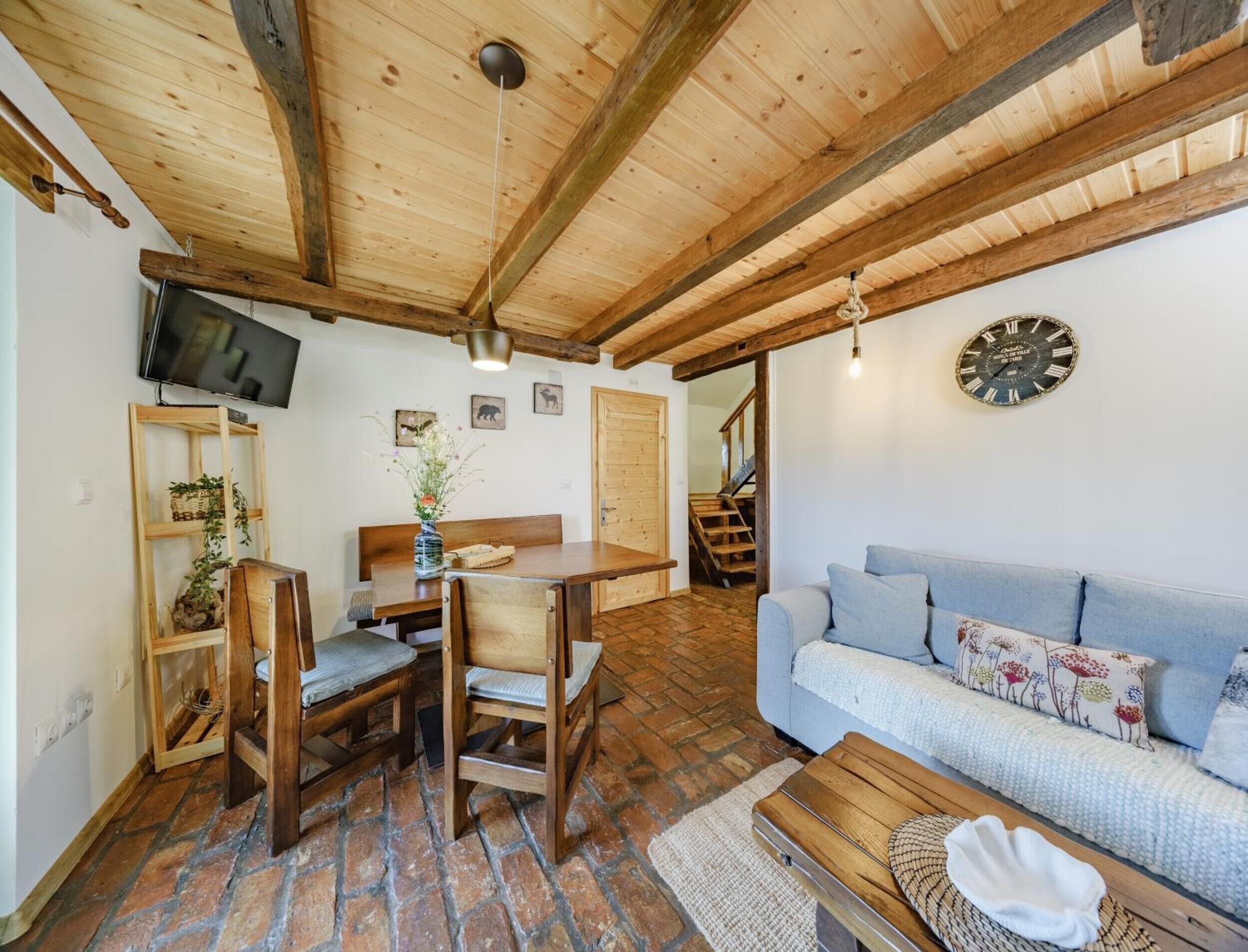 Una sala de estar acogedora con techo de madera, un sofá, una mesa de comedor con sillas, un televisor montado en la pared y un reloj.