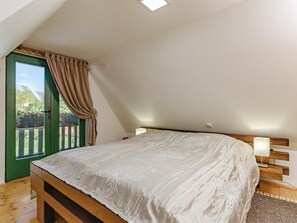 Une chambre avec un plafond en pente, un lit recouvert d’une couvre-lit à motifs floraux, une tête de lit en bois, une table de chevet avec une lampe, et une vue sur une porte verte et un espace extérieur.