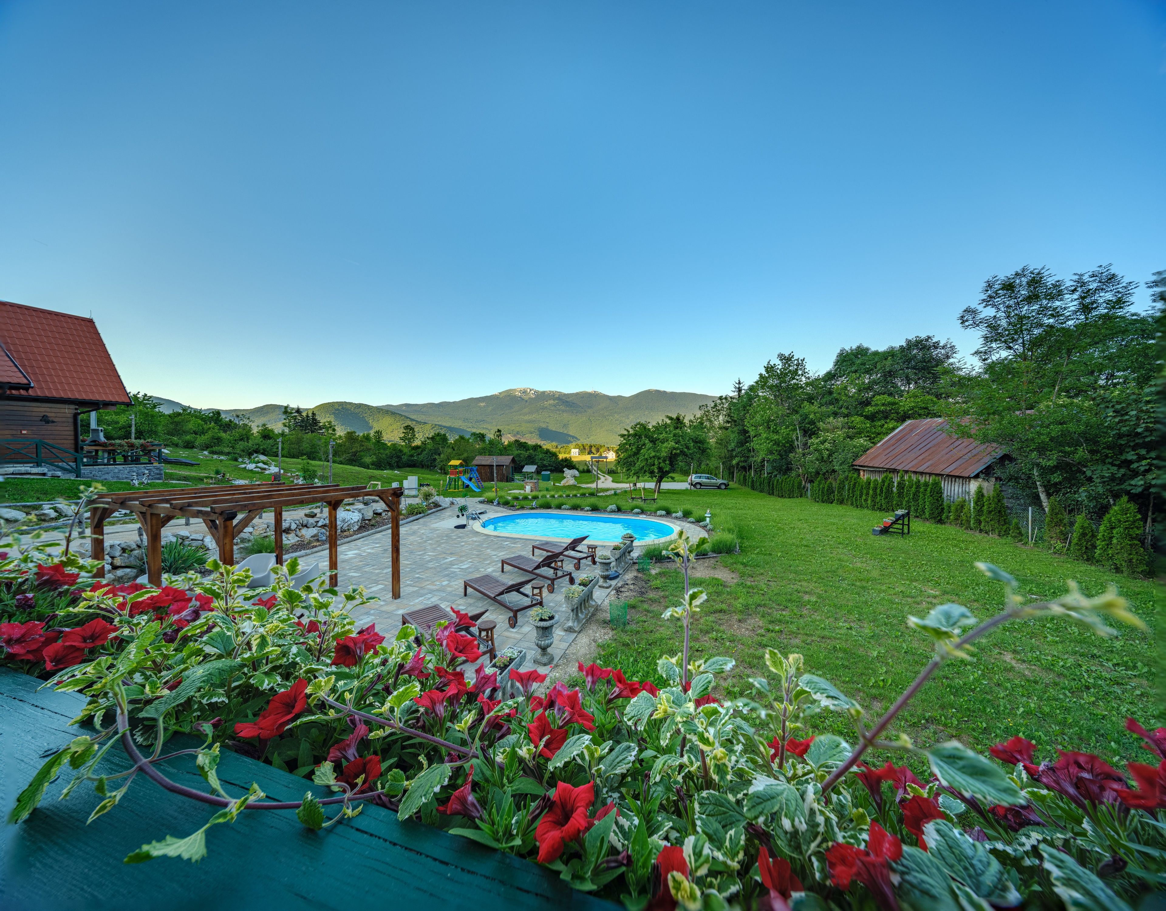 A backyard with a swimming pool, a wooden pergola, and a garden with red flowers.
