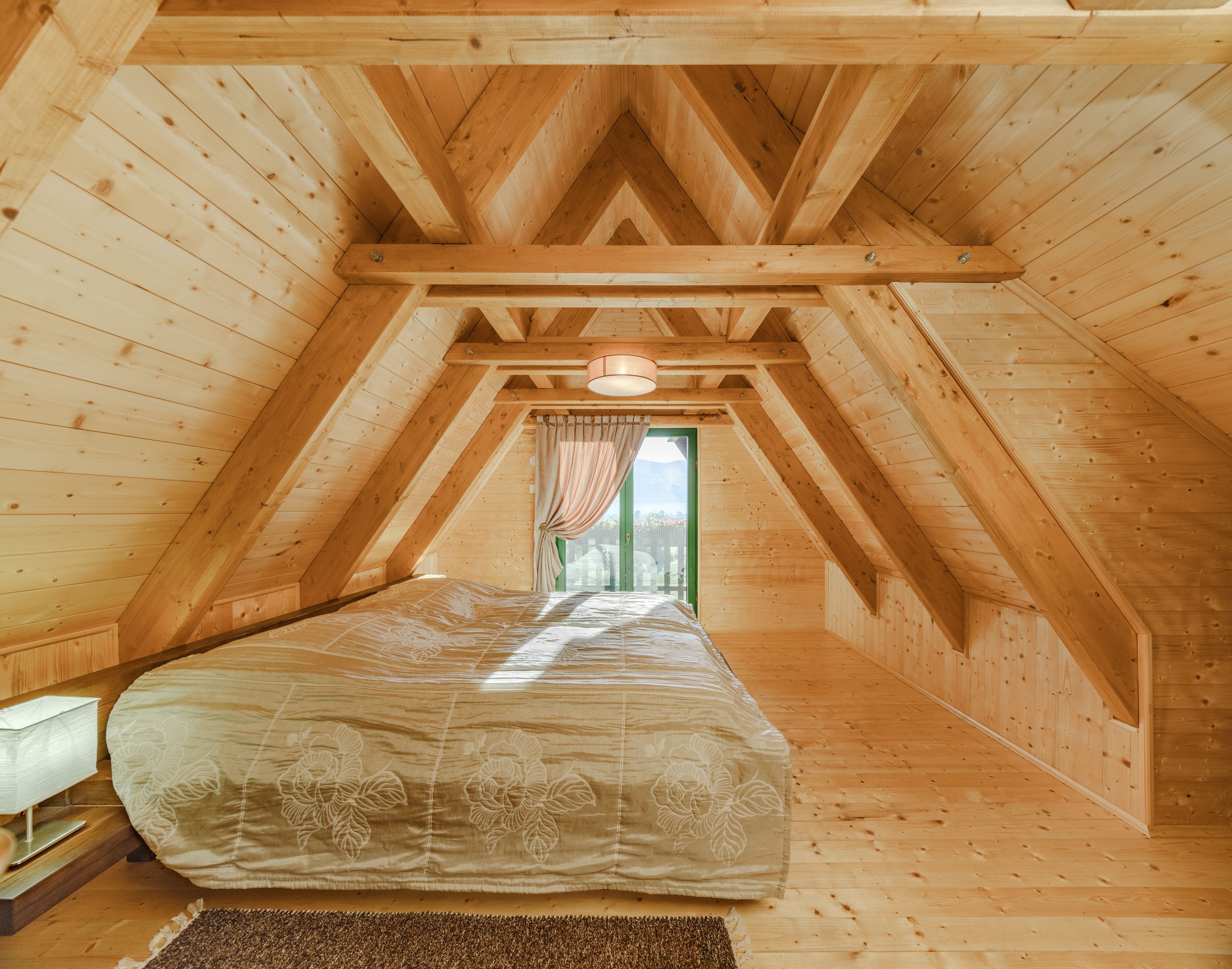 A wooden attic bedroom with a bed, a window with curtains, and a view outside.