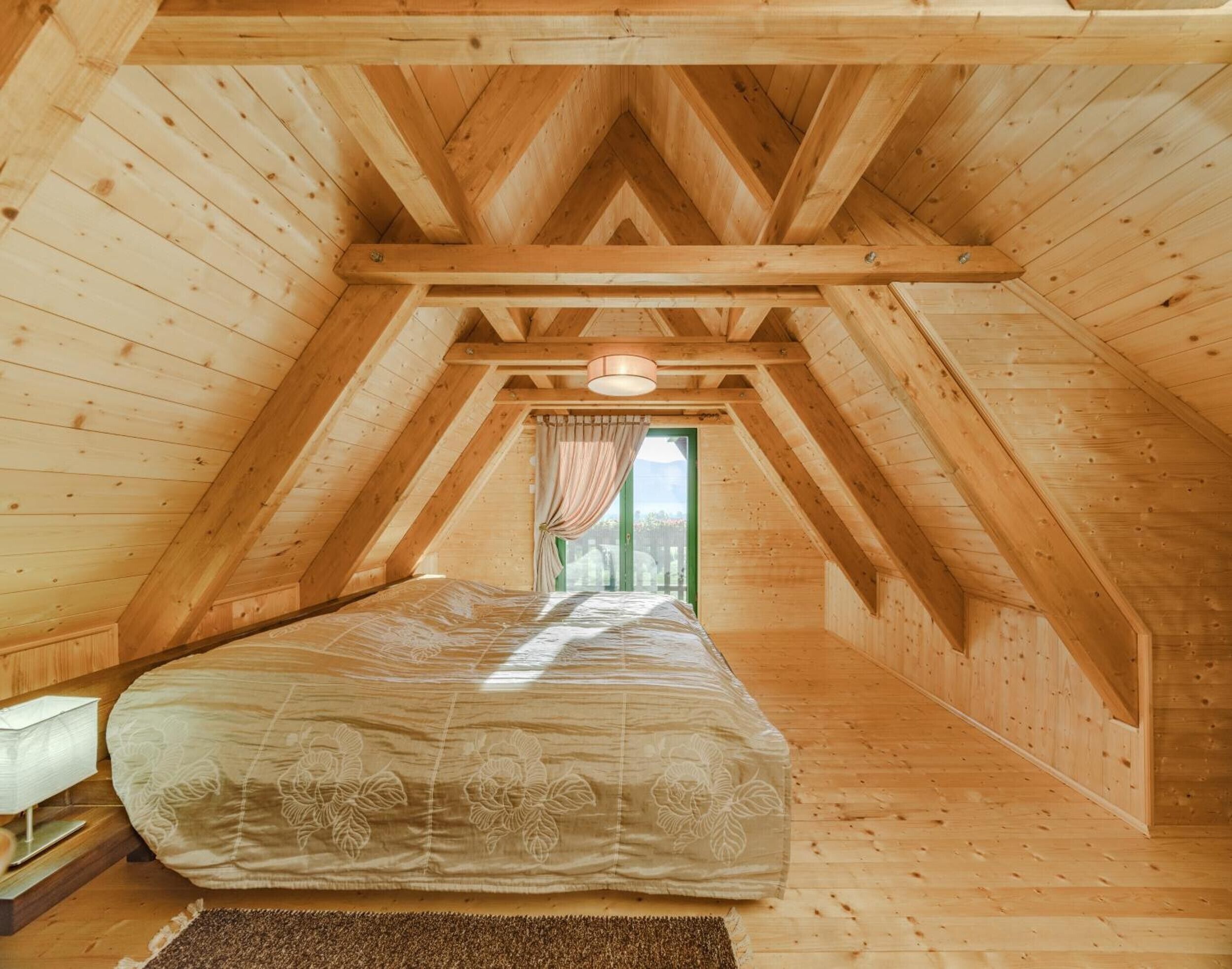 Une chambre avec un plafond en pente, des murs en bois, un lit recouvert d’une couvre-lit à motifs floraux et une fenêtre avec des rideaux.