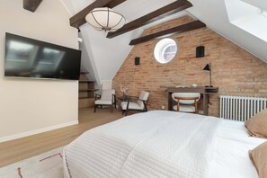 Une chambre avec un plafond en pente, un téléviseur fixé au mur, un lit avec une couette blanche, un bureau avec une chaise et un mur en briques.