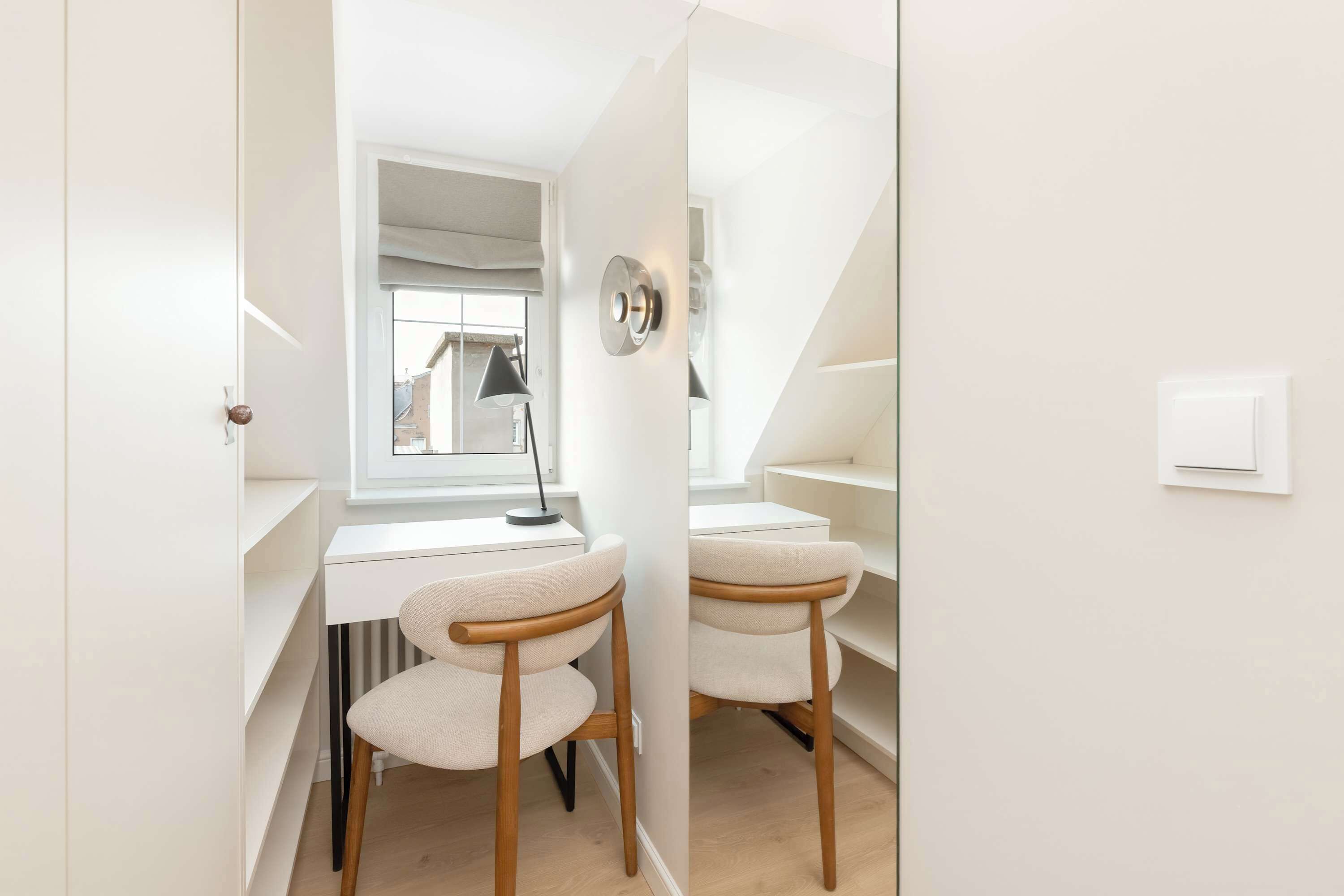 A compact, well-lit room with a desk, two chairs, a window, and a wall-mounted light fixture.