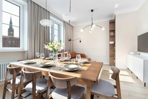 Un coin repas avec une table en bois dressée pour six personnes, entourée de chaises, un vase de fleurs et une vue sur un bâtiment par la fenêtre.