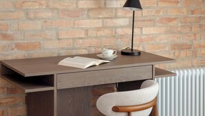 Un bureau en bois, sur lequel se trouvent un livre, une tasse et une lampe, adossé à un mur de briques.
