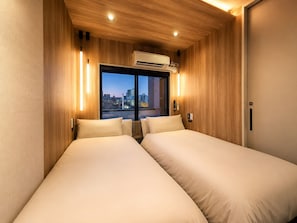 A hotel room with two beds, wooden paneling, an air conditioning unit, a window with a city view, and a door.