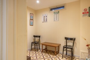 Une petite pièce avec un sol à carreaux, une table en bois, deux chaises et un mur orné de divers panneaux et d’un calendrier.