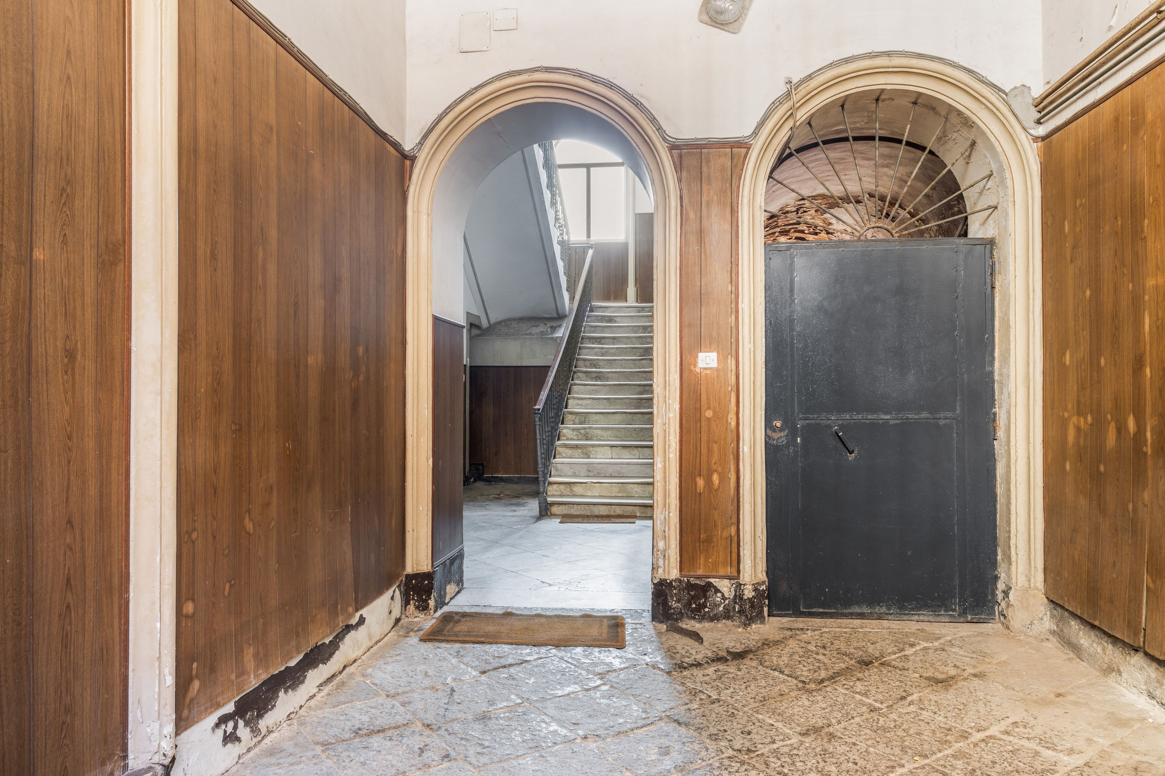 Un pasillo estrecho con paredes de madera y suelo de piedra, que conduce a una escalera y una puerta cerrada.
