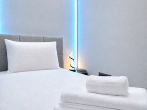 Una cama bien tendida con una almohada y una toalla doblada, una mesita de noche con una lámpara, y una pared con un aplique vertical.