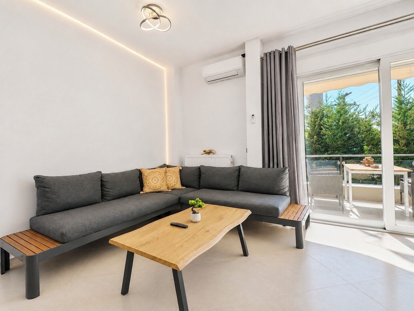 Una sala de estar moderna con un sofá seccional gris, una mesa de centro de madera y un balcón con vista a zonas verdes.