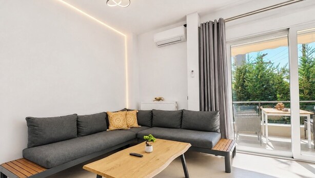 A modern living room with a grey sectional sofa, a wooden coffee table, and a balcony with a view of greenery.