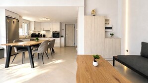 A modern kitchen with white cabinets, a dining area with a wooden table and chairs, and a living area with a sofa and a small plant.