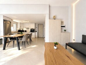 Una cocina moderna con armarios blancos, un comedor con mesa y sillas de madera, y una sala de estar con sofá y una planta pequeña.