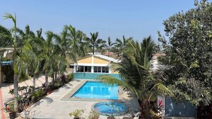 A swimming pool with a surrounding paved area, palm trees, and a building with a yellow roof. - Ocean Blue villa 2bhk (Cheyyur (Taluk) / Vadapattinam)