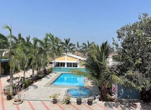 Una piscina con zona pavimentada alrededor, palmeras y un edificio de techo amarillo.